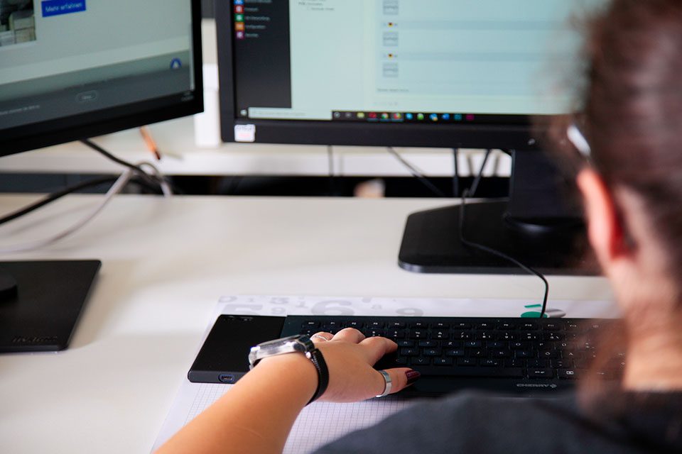 Eine Person mit einer Smartwatch benutzt eine Tastatur an einem Schreibtisch mit zwei Monitoren. Die Bildschirme zeigen verschiedene digitale Inhalte, Details sind jedoch unscharf. Die Umgebung scheint ein Büro oder Arbeitsplatz zu sein.