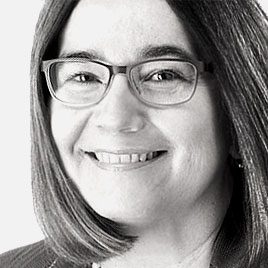 Lächelnde Person mit Brille und schulterlangem Haar, die ein helles Oberteil trägt. Das Bild ist in Schwarzweiß.