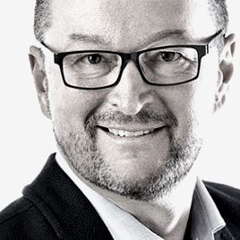 Ein Mann mit kurzen Haaren und Brille lächelt in die Kamera. Er hat einen Bart und trägt ein dunkles Sakko über einem hellen Hemd. Der Hintergrund ist schlicht und hell.