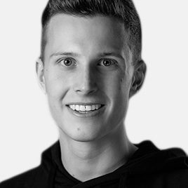Eine Person mit kurzen Haaren lächelt in die Kamera und trägt einen dunklen Kapuzenpullover. Der Hintergrund ist schlicht und hell. Das Bild ist schwarz-weiß.