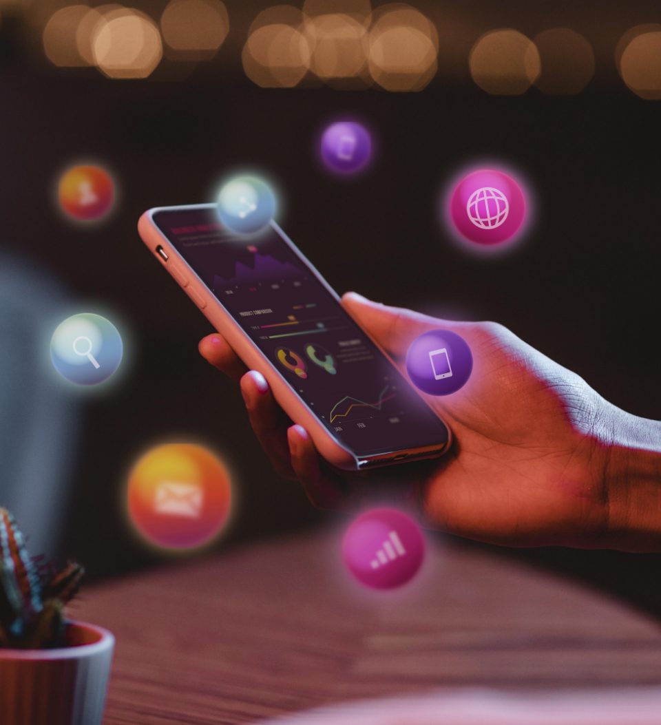 Eine Person hält ein Smartphone, auf dem verschiedene bunte digitale Symbole schweben, darunter ein Globus, eine Lupe und ein Umschlag. Der Hintergrund ist leicht verschwommen und suggeriert Technologie und Konnektivität in einem modernen Umfeld.