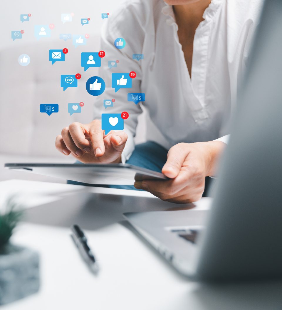Eine Person nutzt ein Tablet, auf dem verschiedene Social-Media-Benachrichtigungssymbole wie „Gefällt mir“, „Herzen“ und „Nachrichten“ schweben, die digitale Interaktion und Konnektivität symbolisieren. Auf einem Schreibtisch in der Nähe sind ein Stift und ein Laptop zu sehen.