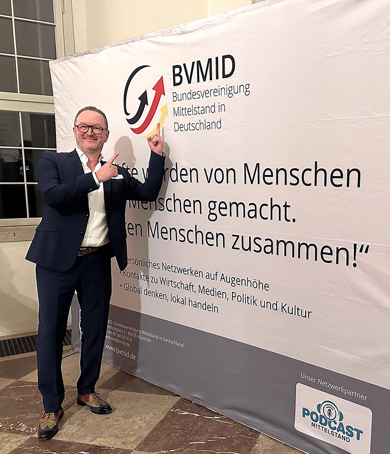 Ein Mann im Anzug lächelt und zeigt auf ein großes Banner. Das Banner trägt das Logo und den Text der BVMID (Bundesvereinigung Mittelstand in Deutschland) und wirbt für Vernetzung und Zusammenarbeit.