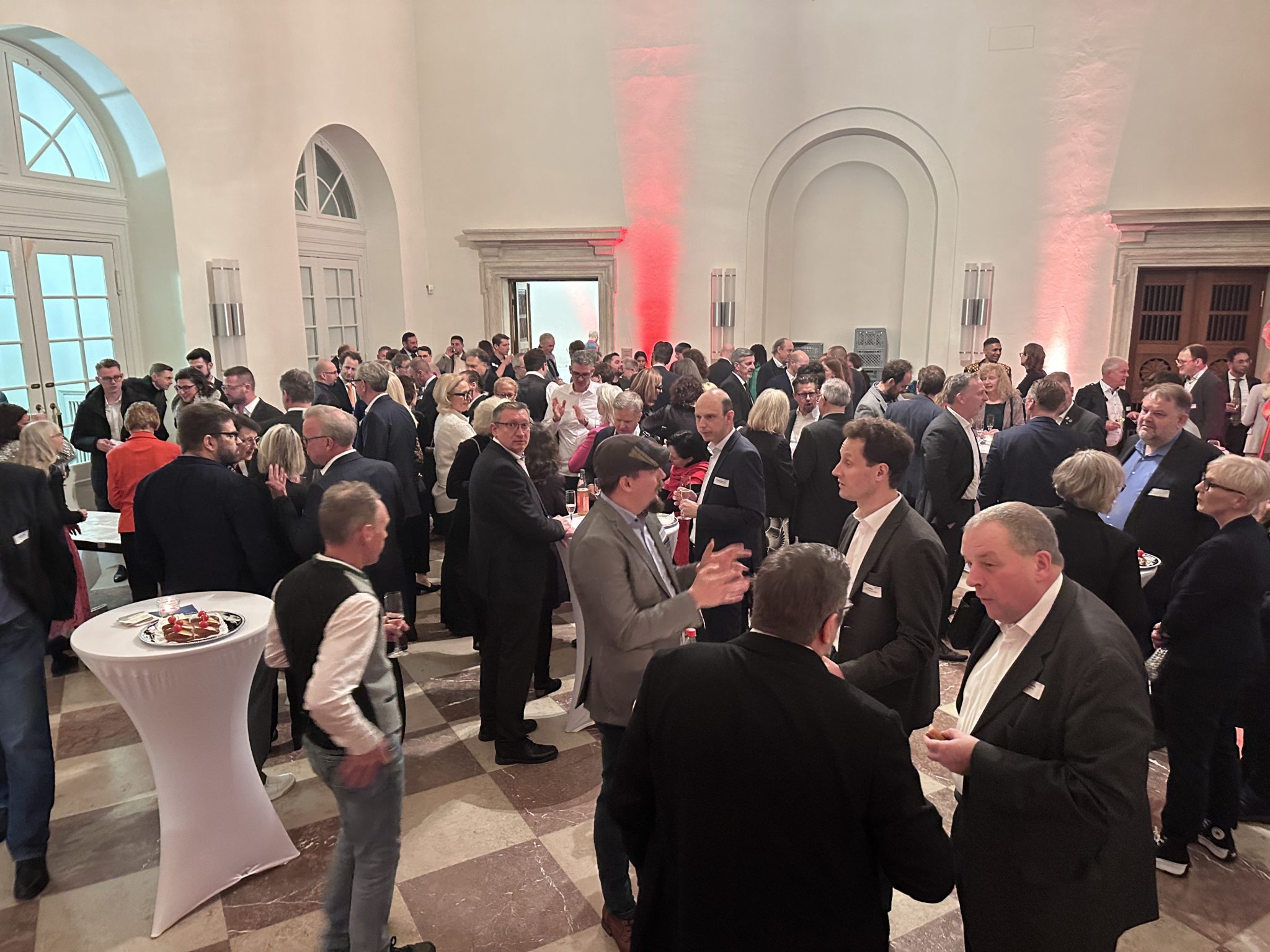 Eine große Gruppe von Menschen in Businesskleidung trifft sich bei einer Networking-Veranstaltung in einem geräumigen, hellen Raum. Sie sitzen an mehreren Tischen mit Erfrischungen. Der Raum hat Bogenfenster und eine hohe Decke.
