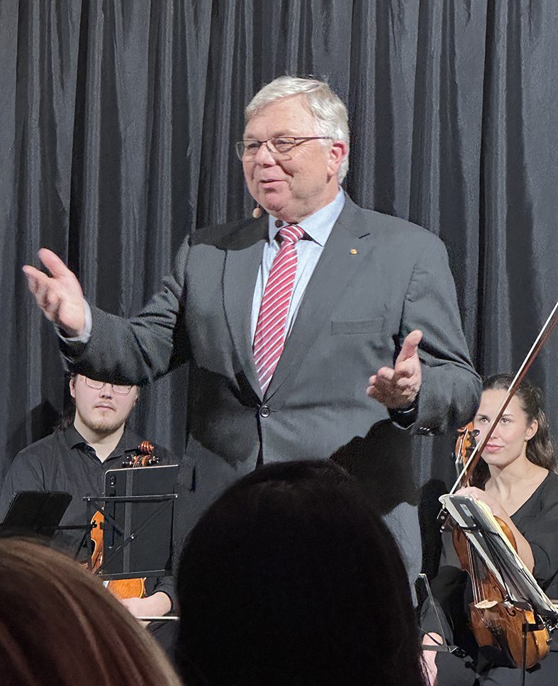 Ein älterer Mann in Anzug und gestreifter Krawatte spricht energisch auf der Bühne. Hinter ihm sitzen eine Frau mit einem Cello und ein Mann mit einer Geige, was mit den schwarzen Vorhängen im Hintergrund an eine musikalische Aufführung erinnert.