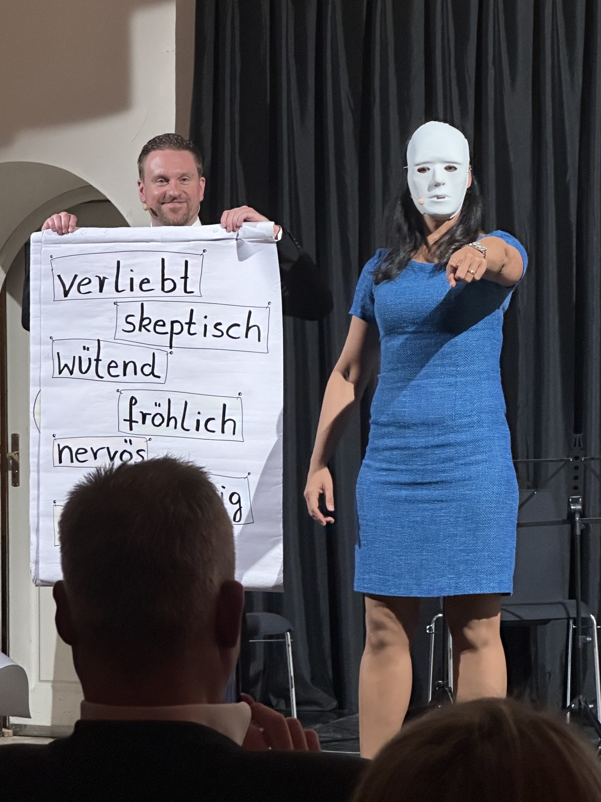 Eine Person in einem blauen Kleid trägt eine weiße Maske und zeigt nach vorne. Eine andere Person steht dahinter und hält ein Plakat mit deutschen Wörtern wie „verliebt“ und „wütend“. Sie stehen auf der Bühne vor einem sitzenden Publikum.
