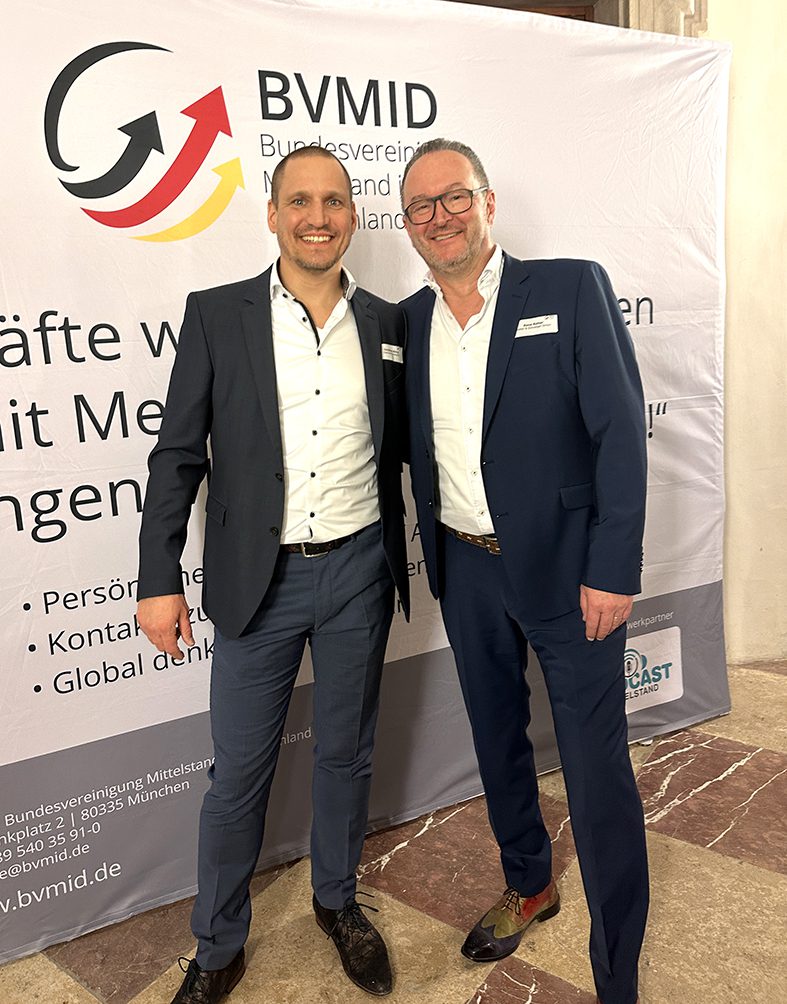 Zwei Männer in Anzügen stehen lächelnd vor einem Banner mit dem BVMID-Logo. Sie befinden sich in einem Gebäude mit Marmorboden. Beide scheinen an einer beruflichen Veranstaltung oder Versammlung teilzunehmen.