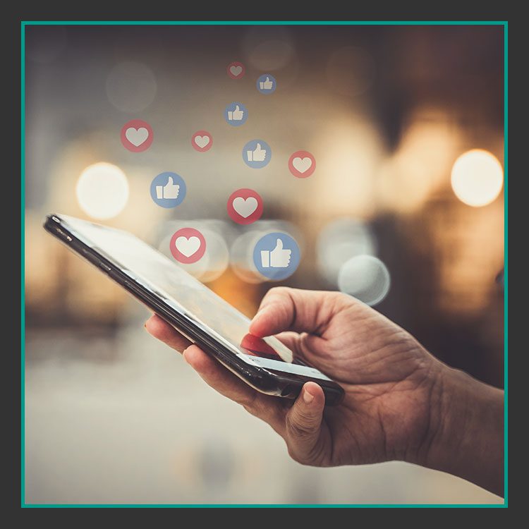 Eine Hand hält ein Smartphone mit erhobenem Daumen und schwebenden Herzsymbolen, die Social-Media-Reaktionen symbolisieren. Der Hintergrund ist durch warmes Licht verschwommen und schafft so eine konzentrierte und zugleich lebendige Atmosphäre.