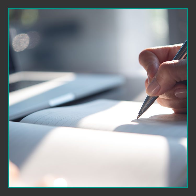 Nahaufnahme einer Hand, die einen Stift hält und in ein Notizbuch schreibt. Die Szene ist gut beleuchtet, Sonnenlicht wirft weiche Schatten auf die Seiten. Im Hintergrund sind ein unscharfer Laptop und ein weiteres Notizbuch zu sehen.