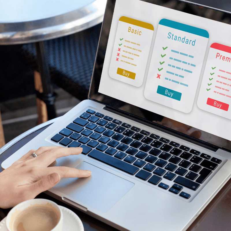 Eine Person sitzt mit einem Laptop an einem Cafétisch im Freien. Auf dem Bildschirm werden drei Preismodelle angezeigt: Basic, Standard und Premium, jeweils mit Checklisten und Kauf-Buttons. Neben dem Laptop steht eine Tasse Kaffee.