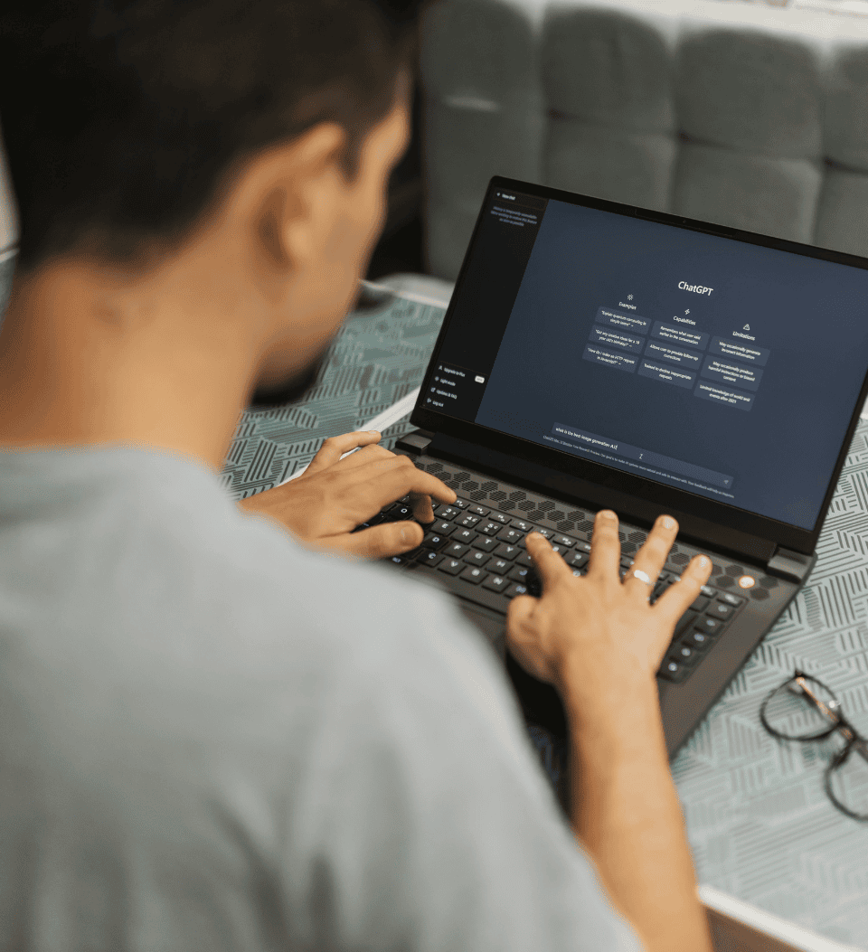 Eine Person tippt auf einem Laptop, der die ChatGPT-Schnittstelle anzeigt. Der Laptop steht auf einem gemusterten Tisch, daneben liegt eine Brille. Das Bild ist auf den Laptop und die Hände fokussiert, wobei das Gesicht der Person nicht zu sehen ist.