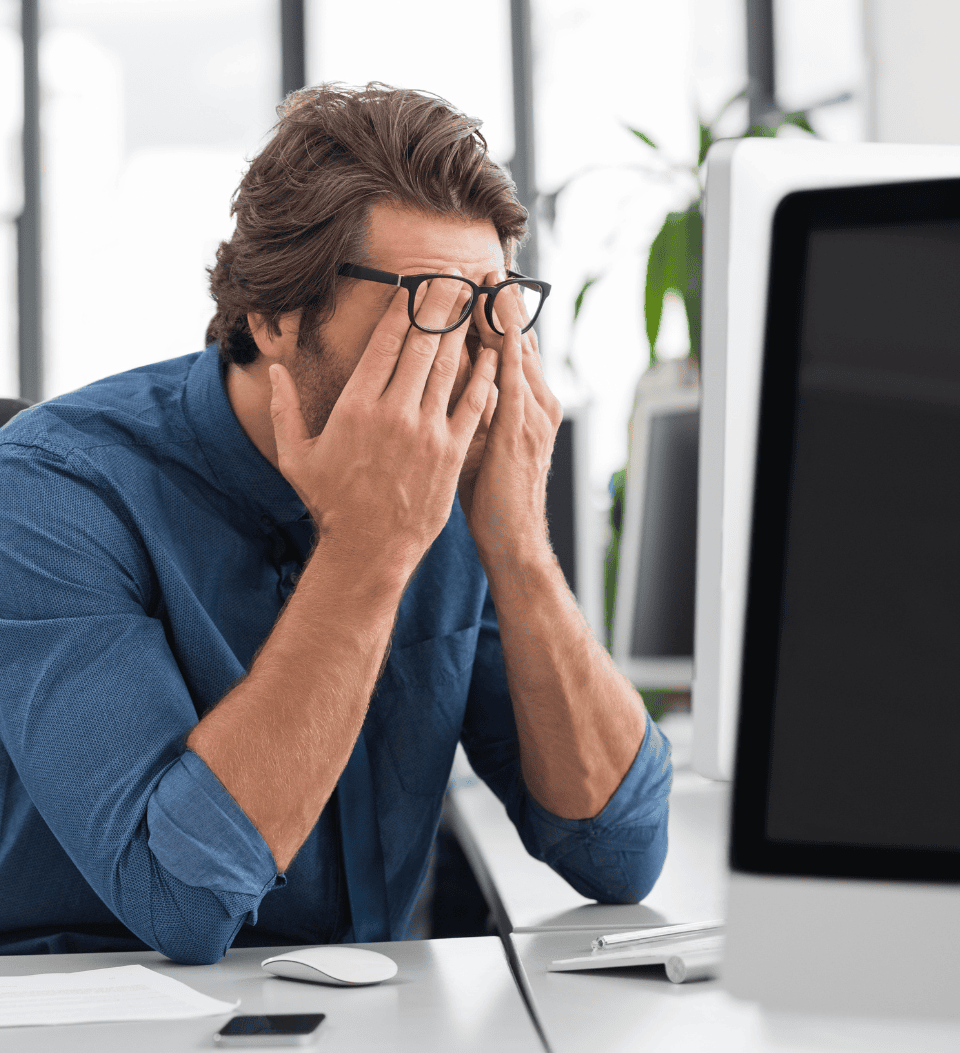 Ein Mann, der an einem Schreibtisch vor einem Computer sitzt, eine Brille und ein blaues Hemd trägt, die Hände vor dem Gesicht verschränkt und in einer Büroumgebung gestresst oder müde wirkt.