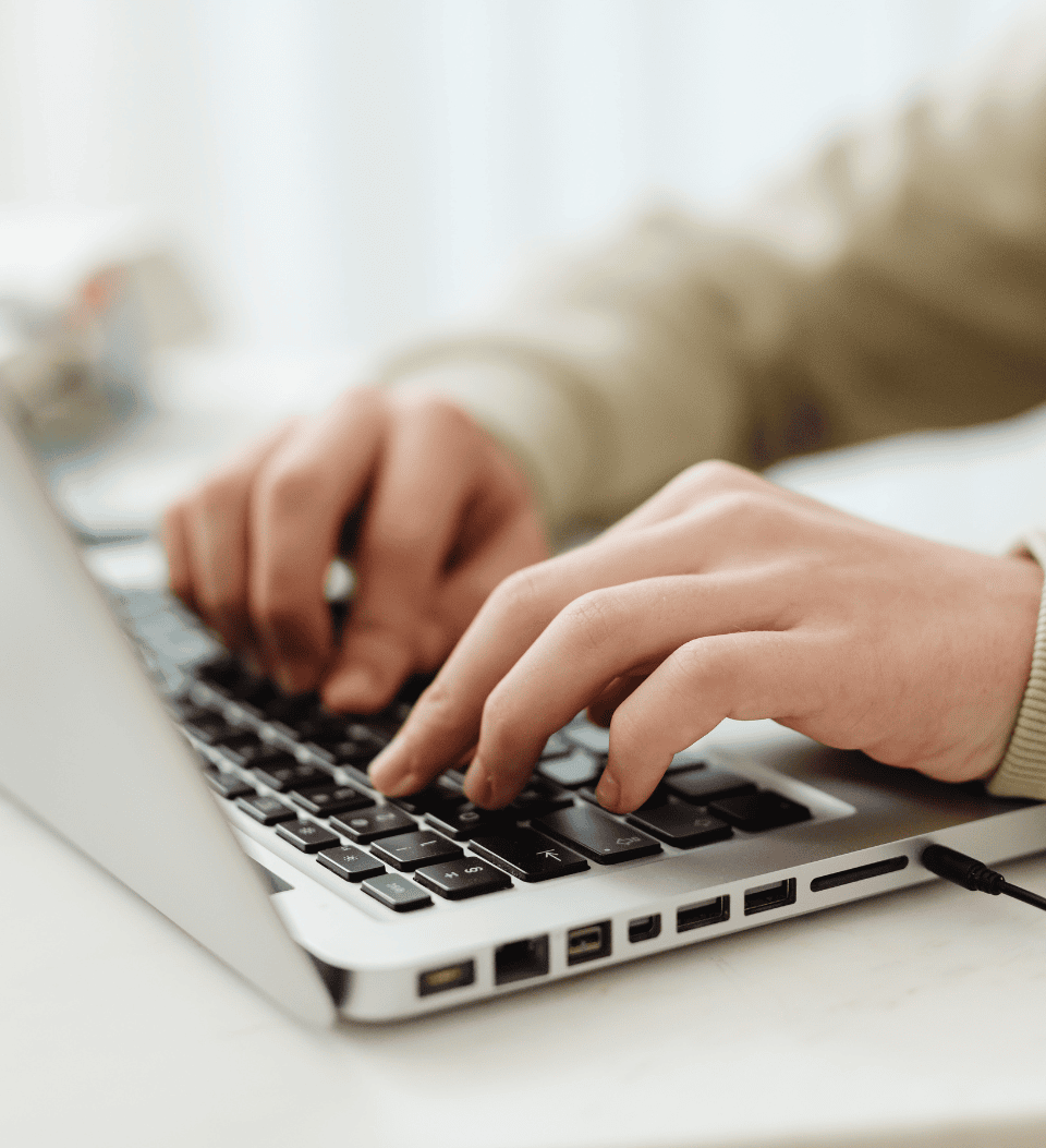 Nahaufnahme der Hände einer Person, die auf einer Laptoptastatur tippen, wobei der Laptop auf einer weißen Fläche steht. Der Hintergrund ist sanft verschwommen.