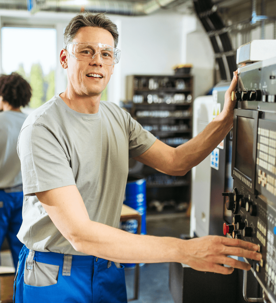 Ein Mann mit Schutzbrille und grauem Hemd bedient ein großes Bedienfeld oder eine Industriemaschine in einer Werkstatt oder Fabrik. Im Hintergrund sind Regale und ein weiterer Arbeiter zu sehen.