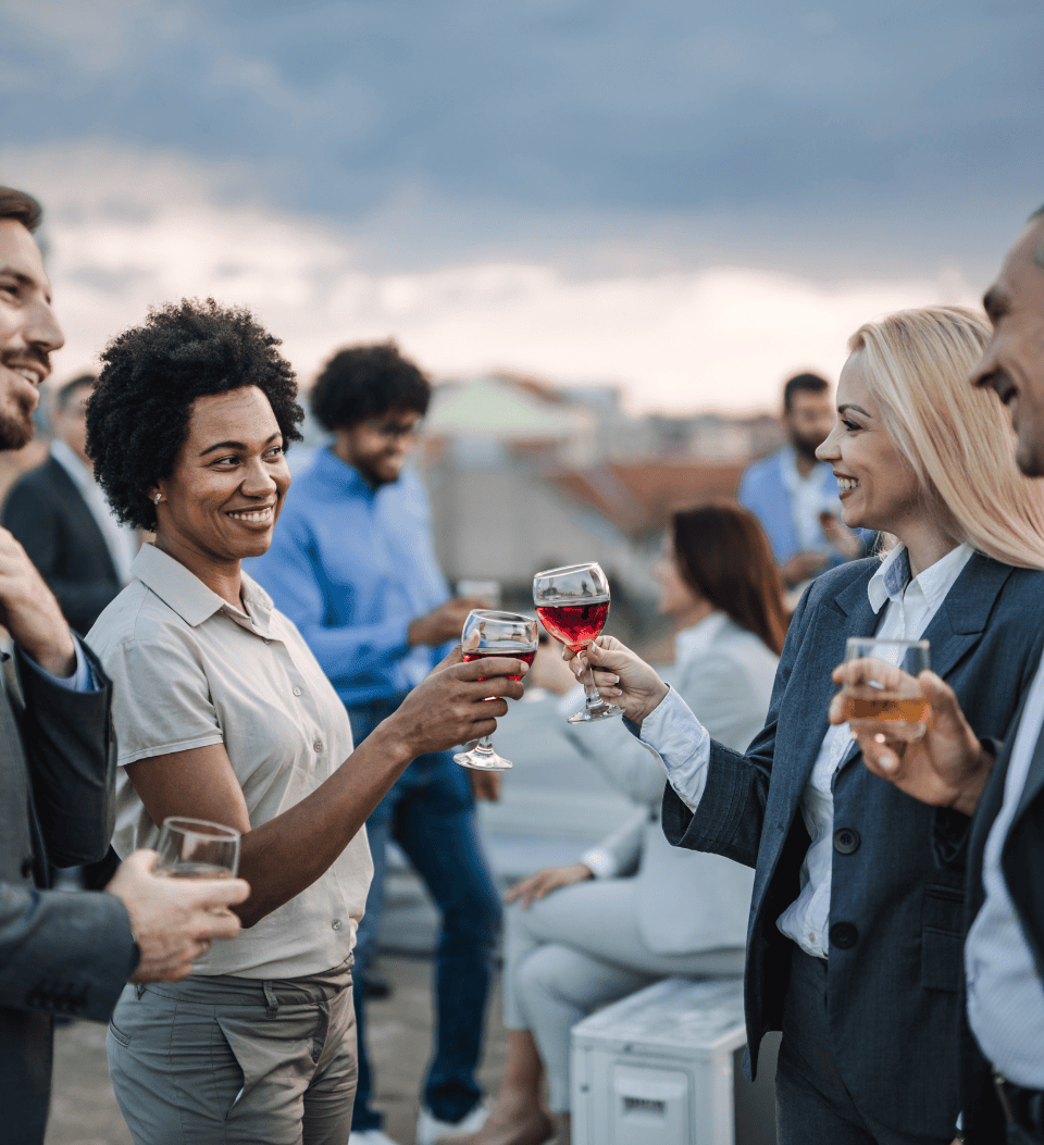 Eine Gruppe von Fachleuten steht im Freien, lächelt und stößt mit Weingläsern an, mit einem bewölkten Himmel im Hintergrund. Zwei Frauen im Vordergrund stoßen mit ihren Gläsern an, während andere in der Nähe plaudern und sich unter die Leute mischen.