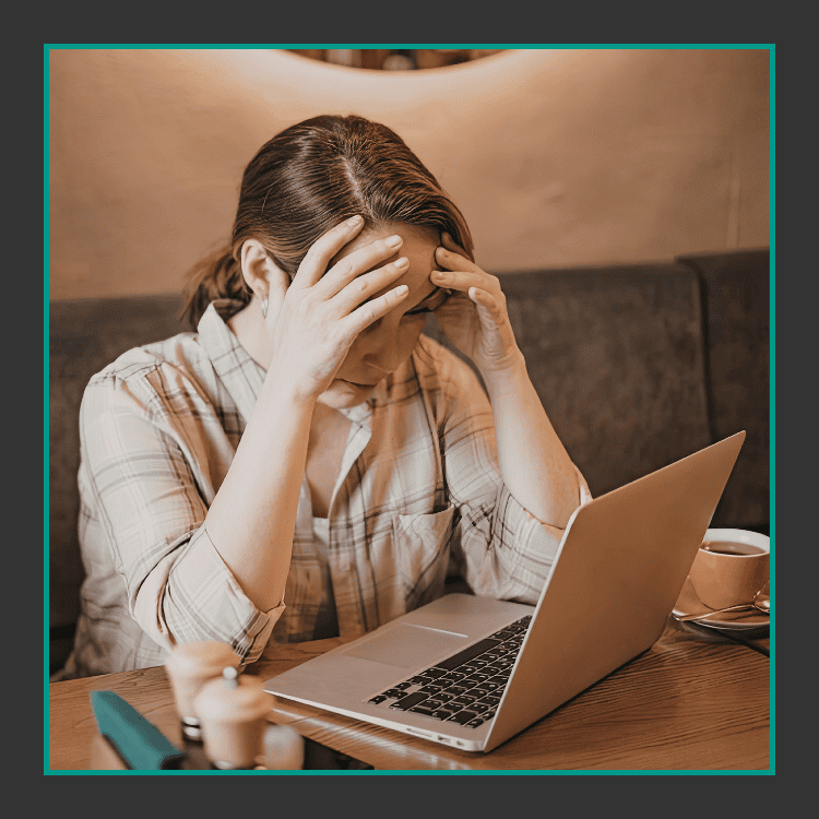 Eine Frau sitzt an einem Tisch mit einem Laptop, hält ihren Kopf in den Händen und wirkt gestresst oder frustriert. Auf dem Tisch stehen eine Tasse Kaffee und einige Gegenstände, und sie befindet sich im Haus.