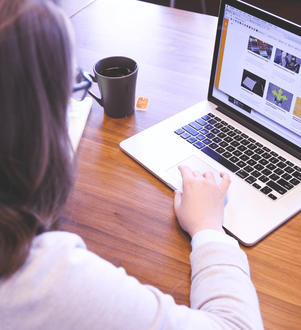 Eine Person mit schulterlangem Haar arbeitet mit einem Laptop an einem Holztisch. Eine Tasse und ein Teebeutel stehen daneben. Auf dem Bildschirm des Laptops wird eine Website mit Bildern und Artikeln angezeigt.