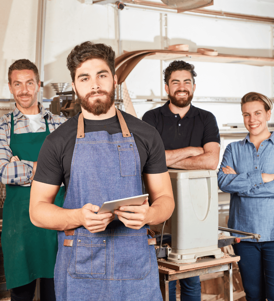 Vier Personen in einer Werkstatt: Ein Mann in einer blauen Schürze hält eine Tafel, ein Mann in einer grünen Schürze steht links von ihm, und zwei lächelnde Personen stehen hinter ihnen.