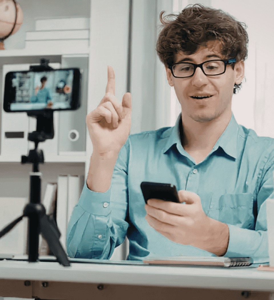 Ein Mann mit Brille und blauem Hemd sitzt an einem Schreibtisch, hält ein Smartphone in der Hand und gestikuliert mit erhobenem Finger, während er sich selbst mit einem Telefon auf einem Stativ aufnimmt. Im Hintergrund sind Regale mit Büchern zu sehen.