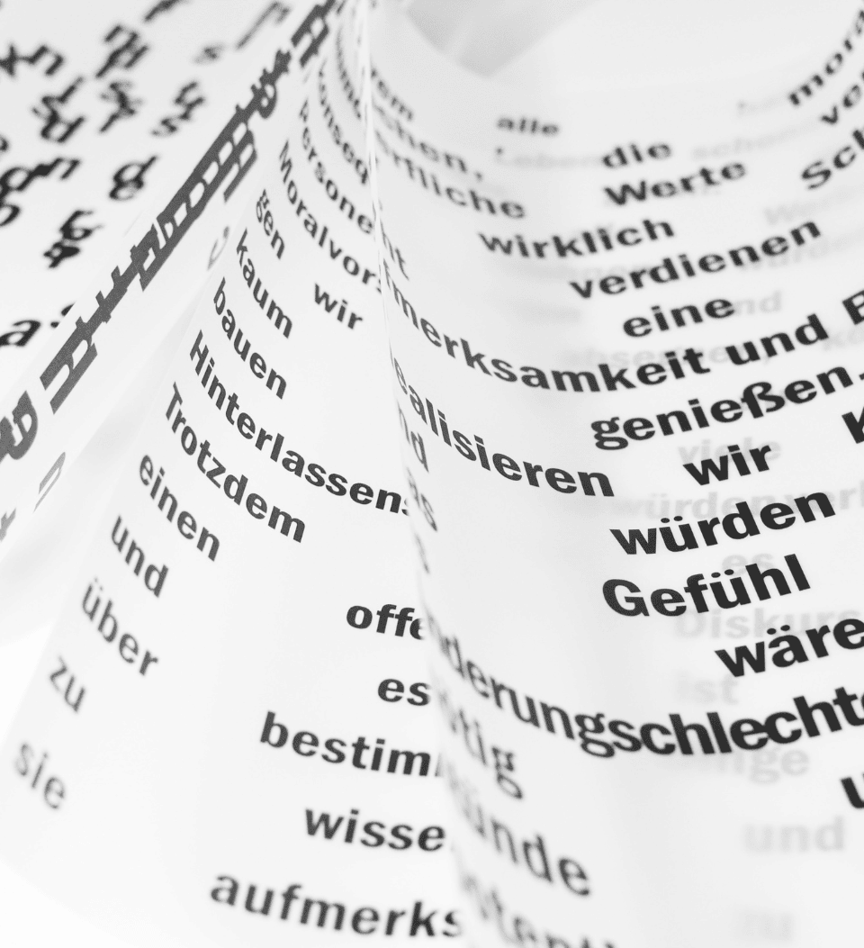 Eine Nahaufnahme von weißen Blättern, die mit verstreuten schwarzen deutschen Wörtern und Sätzen bedruckt sind, einschließlich Elementen von Website-Texte, die kunstvoll angeordnet und leicht gewellt sind, um einen abstrakten, geschichteten visuellen Effekt zu erzeugen.