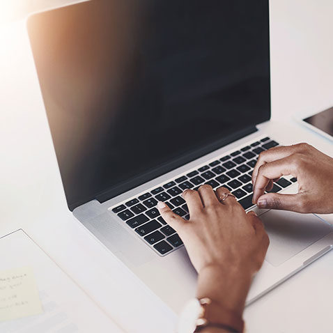 Eine Person tippt auf einer Laptoptastatur mit leerem Bildschirm, umgeben von Papieren und einem Smartphone. Sonnenlicht beleuchtet die Szene von der oberen linken Ecke.