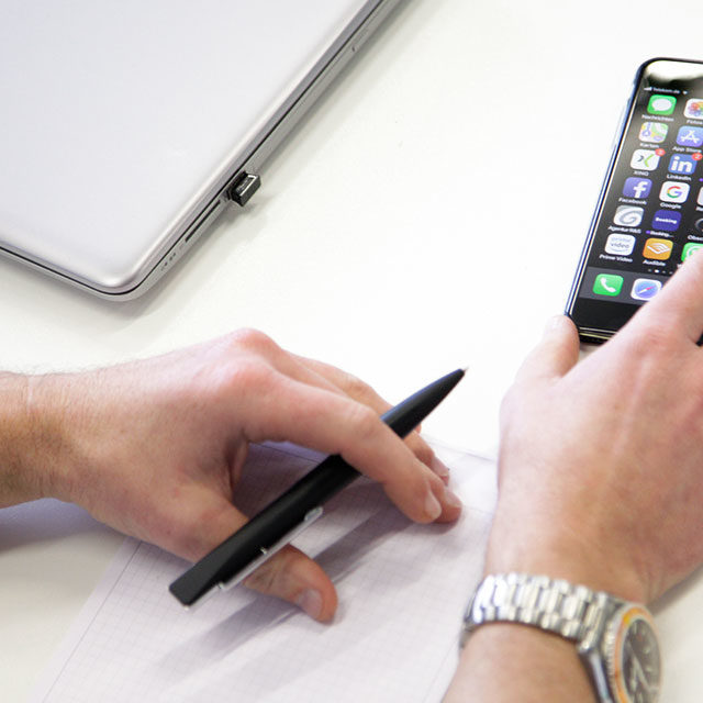 Eine Person hält einen Stift über einen leeren Notizblock. Daneben liegen ein Smartphone und ein Laptop auf einem weißen Schreibtisch. Auf dem Display des Telefons werden verschiedene App-Symbole angezeigt. Die Person trägt eine Uhr am linken Handgelenk.