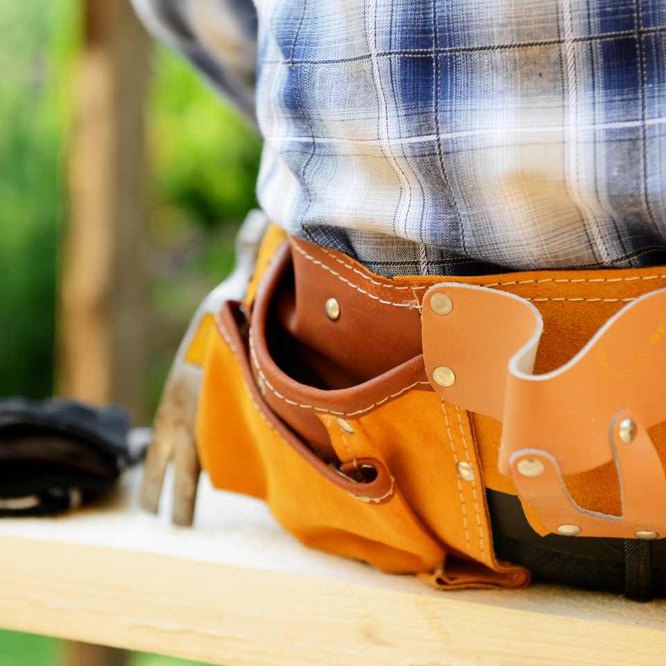 Nahaufnahme einer Person in einem blau karierten Hemd, die einen ledernen Werkzeuggürtel mit einem Hammer und Werkzeugen trägt und an einer hölzernen Arbeitsfläche im Freien steht.