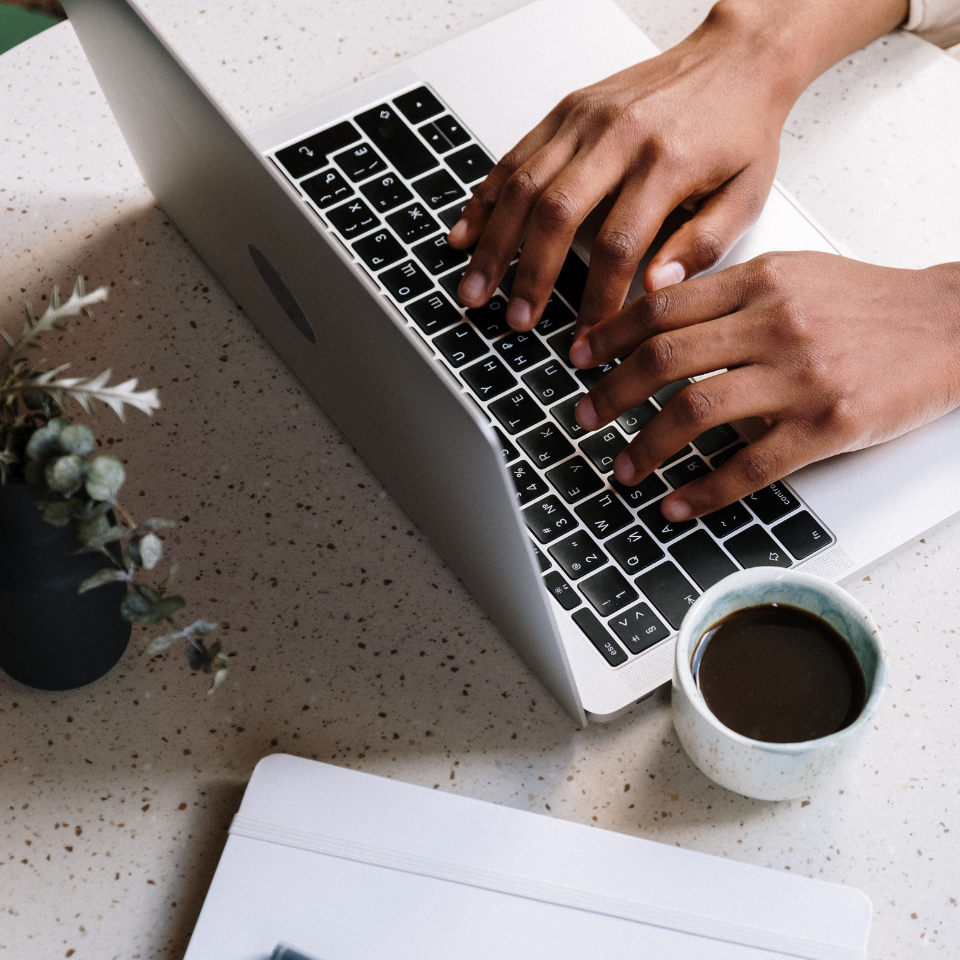Eine Person tippt auf einem Laptop an einem gesprenkelten Tisch mit einer Tasse Kaffee, einer kleinen Topfpflanze und einem geschlossenen Notizbuch mit einem Stift in der Nähe und fängt damit die Essenz der Stock-Medien-Arbeitsplatzinspiration ein.