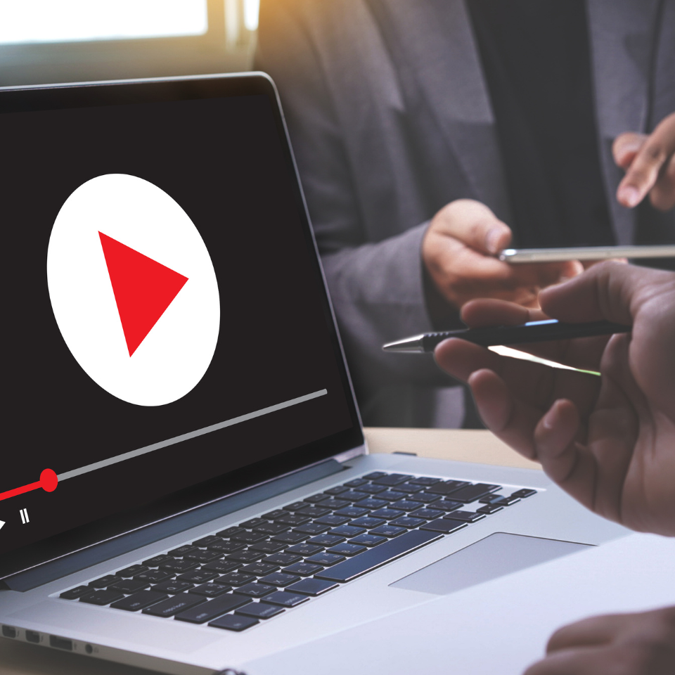 Nahaufnahme eines Laptops mit einem Videoplayer und einer großen Wiedergabetaste auf dem Bildschirm. In der Nähe sind zwei Hände zu sehen, von denen eine einen Stift und die andere ein Tablet hält, was auf eine Diskussion oder Besprechung hindeutet.
