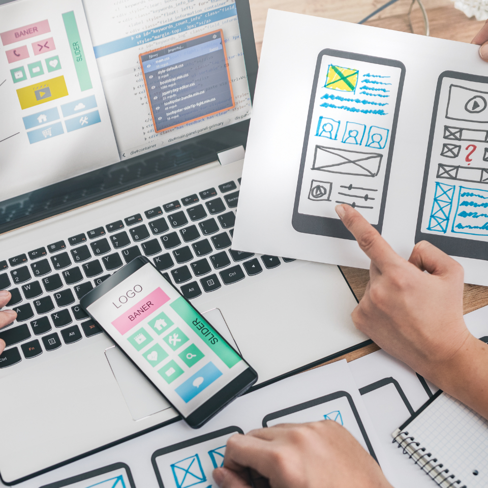 Menschen, die gemeinsam an der Entwicklung einer mobilen App arbeiten, mit Wireframe-Skizzen auf Papier, einem Smartphone und einem Laptop, auf dem Schnittstellenlayouts und Code angezeigt werden. Die Hände zeigen auf verschiedene Designelemente, was auf eine Brainstorming-Sitzung hindeutet.